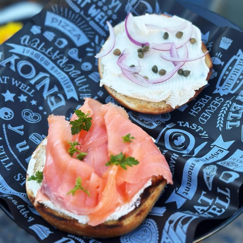 Two bagels on black paper, one with salmon and parsley, the other with cream cheese, red onion, and capers.