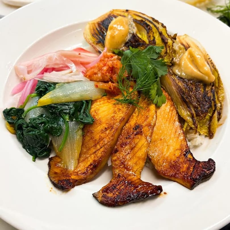 A white plate with grilled fish, bok choy, kimchi, pickled onions, and charred cabbage.