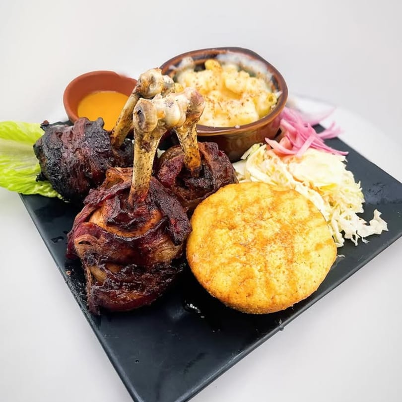 A plate of bacon-wrapped chicken drumsticks, mac and cheese, coleslaw, cornbread, and dipping sauce.