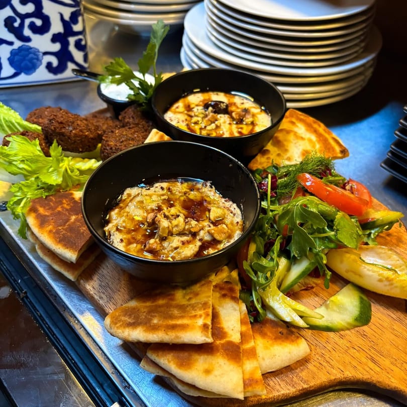 A wooden board with falafel, pita bread, two bowls of hummus, and a side salad.