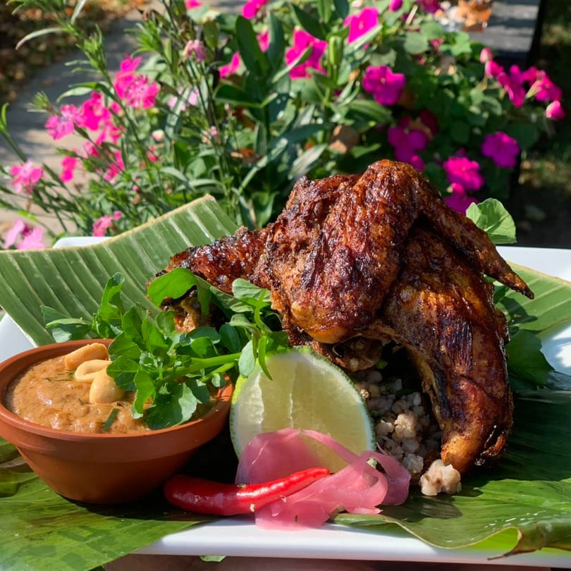 Grilled chicken with peanut sauce, lime, and pickled onions on a banana leaf, with pink flowers in the background.
