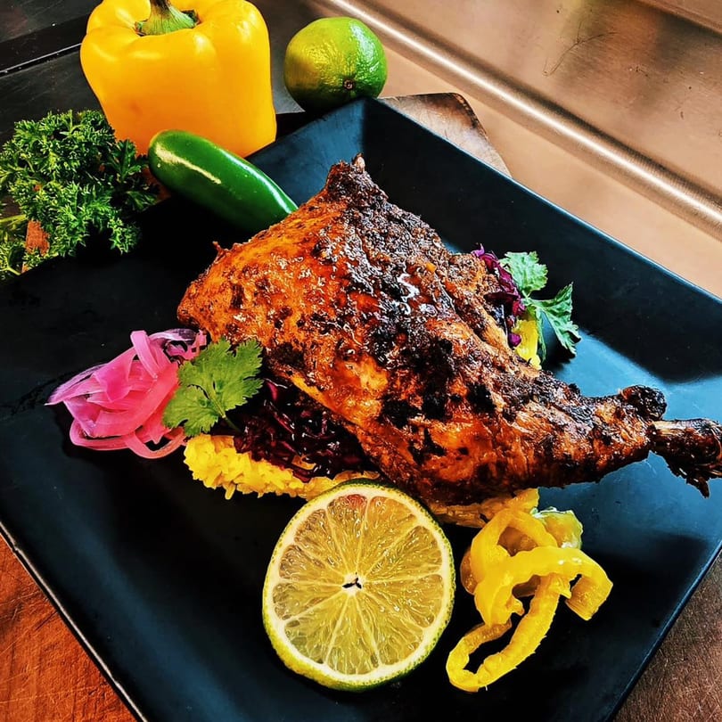 Spicy roasted chicken leg and thigh with yellow rice, pickled onions, and lime on a black plate.
