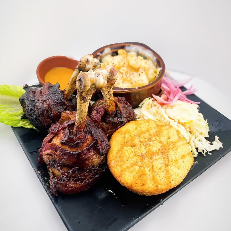 A plate of bacon-wrapped chicken drumsticks, mac and cheese, cornbread, coleslaw, and dipping sauce.