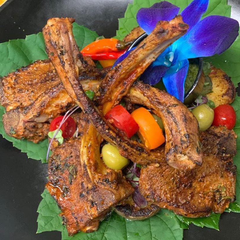 Grilled lamb chops on green leaves with colorful peppers, olives, and a purple orchid garnish.