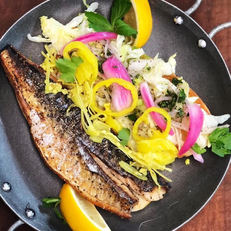 Grilled fish fillet with colorful pickled vegetables, cabbage, and lemon wedges in a dark pan.