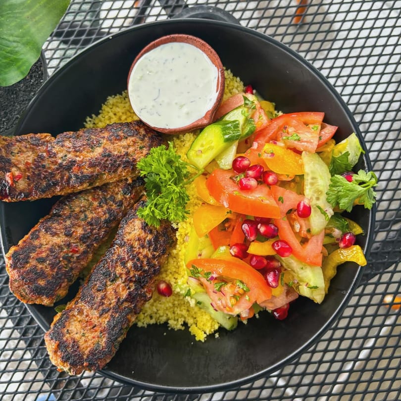 Grilled kofta kebabs with couscous, fresh salad, pomegranate seeds, and a side of white sauce.
