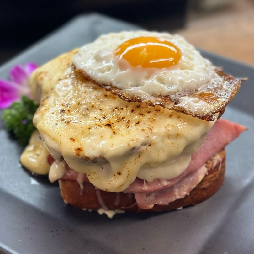 Croque Madame sandwich with melted cheese, ham, and a fried egg on top, garnished with an orchid.