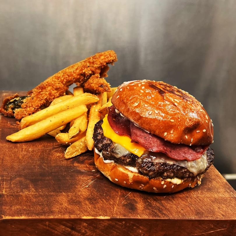 Gourmet burger with melted cheese and pastrami, served with golden fries and fried pickles on a wooden board.