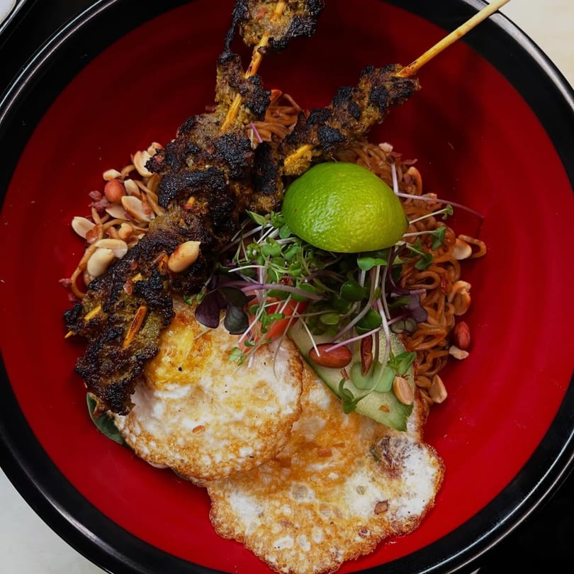 Noodle dish with two fried eggs, two skewers of grilled meat, peanuts, and a lime wedge.