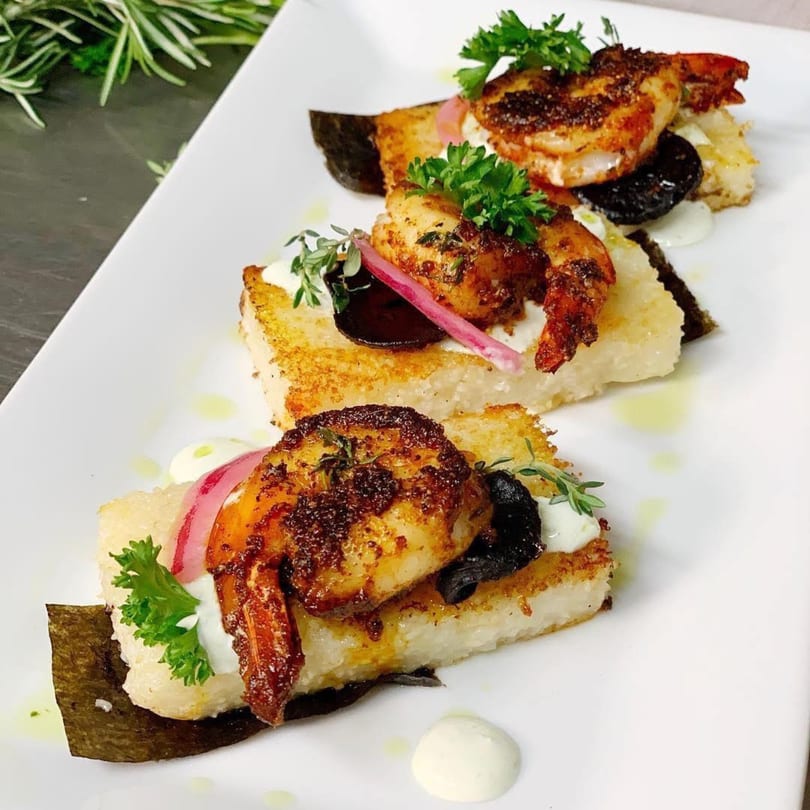 Three shrimp appetizers on a white plate, garnished with herbs, pickled onions, and dark olives.
