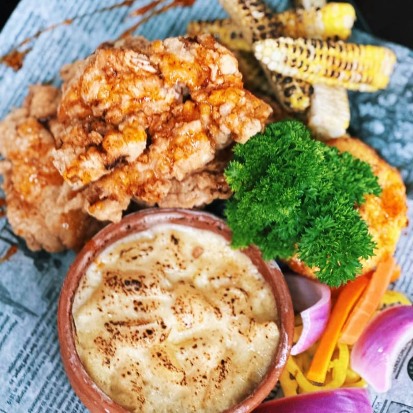 Crispy fried chicken, grilled corn, baked potato, and fresh vegetables on a newspaper-patterned plate.